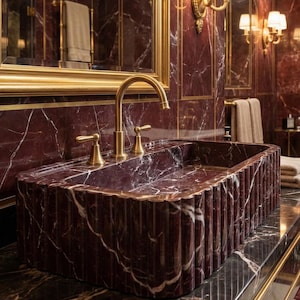 May include: A luxurious bathroom sink made of burgundy marble with white veining. The sink has a fluted design and is paired with a gold faucet and fixtures. The walls are burgundy marble with gold trim, and there is a gold-framed mirror.