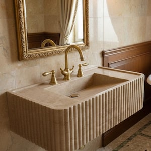May include: A beige, rectangular bathroom sink with a fluted front design. The sink has a gold faucet and handles. A gold-framed mirror hangs above the sink, reflecting the room's details. The sink is mounted on a wall with beige tiles.