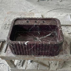 May include: A rectangular, burgundy marble sink with white veining. The sink has a scalloped edge and a decorative, vertical ribbed pattern on the sides. It features three faucet holes and a central drain.