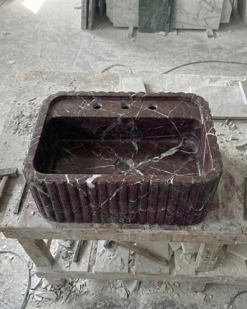 May include: A rectangular, burgundy marble sink with white veining. The sink has a scalloped edge and a decorative, vertical ribbed pattern on the sides. It features three faucet holes and a central drain.