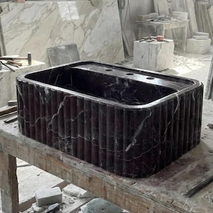 May include: A rectangular, dark burgundy marble sink with vertical fluted sides. The sink has a built-in top with three circular faucet holes. The marble has white veining. The sink is displayed on a wooden workbench in a workshop setting.