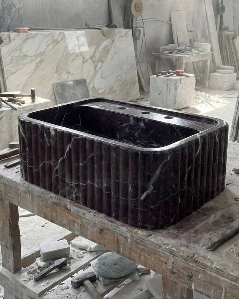 May include: A rectangular, dark burgundy marble sink with vertical fluted sides. The sink has a built-in top with three circular faucet holes. The marble has white veining. The sink is displayed on a wooden workbench in a workshop setting.
