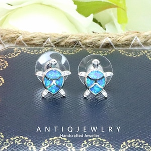 Könnte beinhalten: Silberfarbene Schildkröten-Ohrstecker mit blau schimmernden Opal-Schalen und transparenten Rückseiten. Die Ohrringe werden auf einer dunklen Oberfläche mit dem Text "ANTIQ JEWLRY Handcrafted Jeweller" präsentiert.