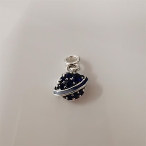 May include: A silver charm featuring a blue planet design with a light blue band. The planet is covered in small, silver-colored dots. The charm has a loop at the top for attaching to a bracelet or necklace.