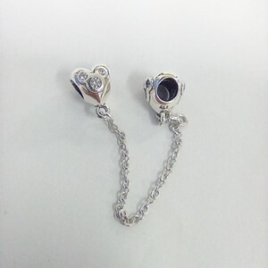 May include: A silver safety chain charm for a bracelet, featuring two cylindrical ends connected by a delicate link chain. One end is shaped like a heart with three embedded clear stones arranged in a circular pattern, while the other is plain.