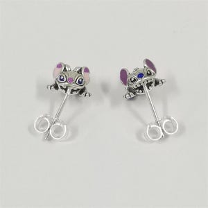 May include: A pair of silver stud earrings featuring a character design with large ears and wide eyes. The character has purple and pink enamel accents on the ears and blue enamel details on the eyes. Each earring includes a silver post and back.