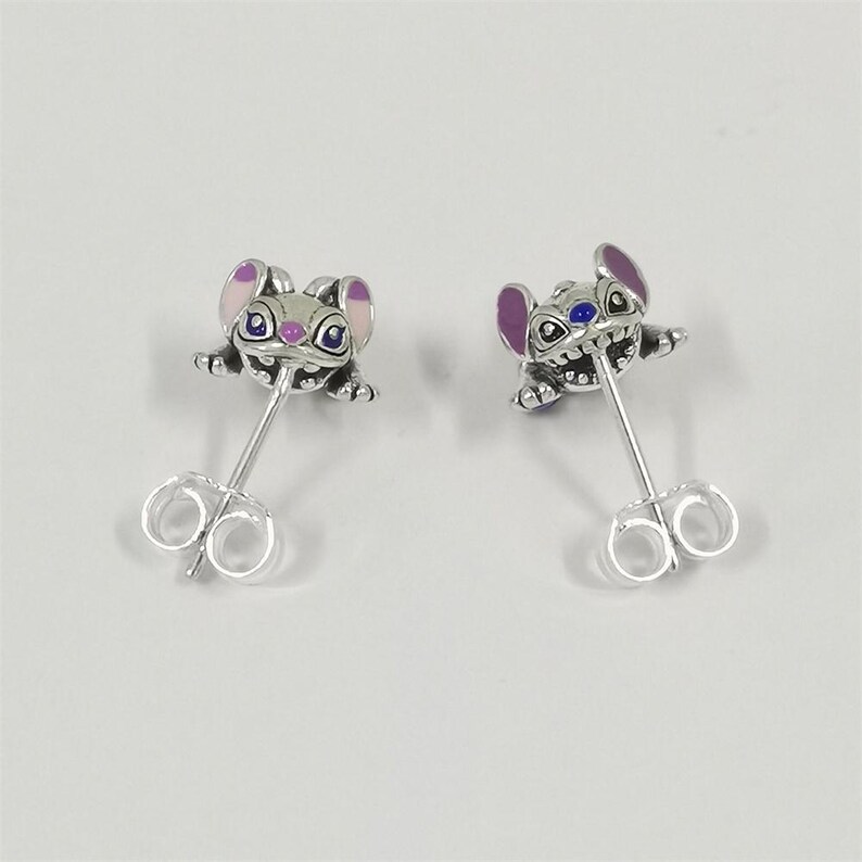 May include: A pair of silver stud earrings featuring a character design with large ears and wide eyes. The character has purple and pink enamel accents on the ears and blue enamel details on the eyes. Each earring includes a silver post and back.