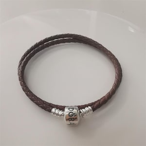 Puede incluir: Una pulsera de cuero marrón con un diseño trenzado. La pulsera presenta un charm plateado en forma de barril con la palabra "Pandora" grabada. La pulsera tiene aproximadamente 18 cm de circunferencia.