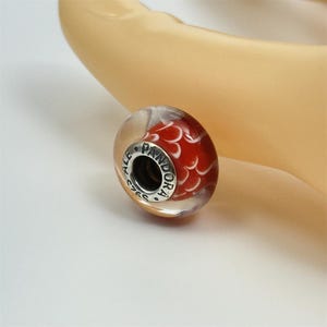 May include: A clear glass bead with a red and white design. The bead has a silver-colored metal core with the word "PANDORA" engraved on it. The bead is round and has a fish scale-like pattern.