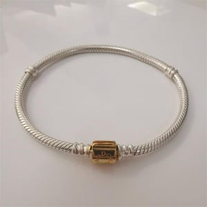 以下が含まれることがあります： シルバーのブレスレット。スネークチェーンのデザイン。 金色のバレルクラスプが付いており、テキストが刻印されています。 ジュエリーです。