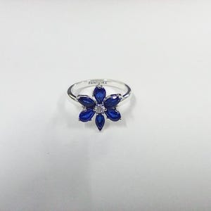 May include: A silver ring featuring a flower-shaped centerpiece composed of six marquise-cut deep blue stones surrounding a single clear center stone. The band is engraved with the word PANDORA on the inner surface, showcasing a polished finish.