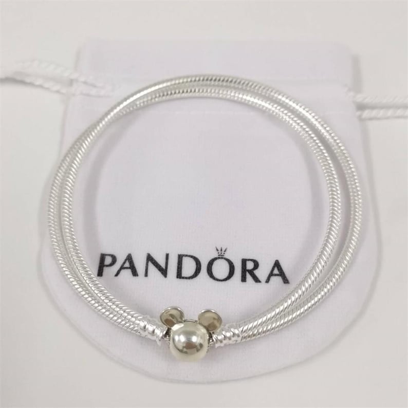 May include: A silver snake chain bracelet featuring a clasp shaped like the head of Mickey Mouse. The bracelet is displayed on a white fabric pouch with the black text PANDORA printed on the front. The metal has a polished, reflective finish.