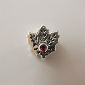 May include: A silver charm in the shape of a maple leaf with intricate detailing. A small, round, red gemstone is set in the center of the leaf. The charm is likely a pendant or bead for a bracelet or necklace.