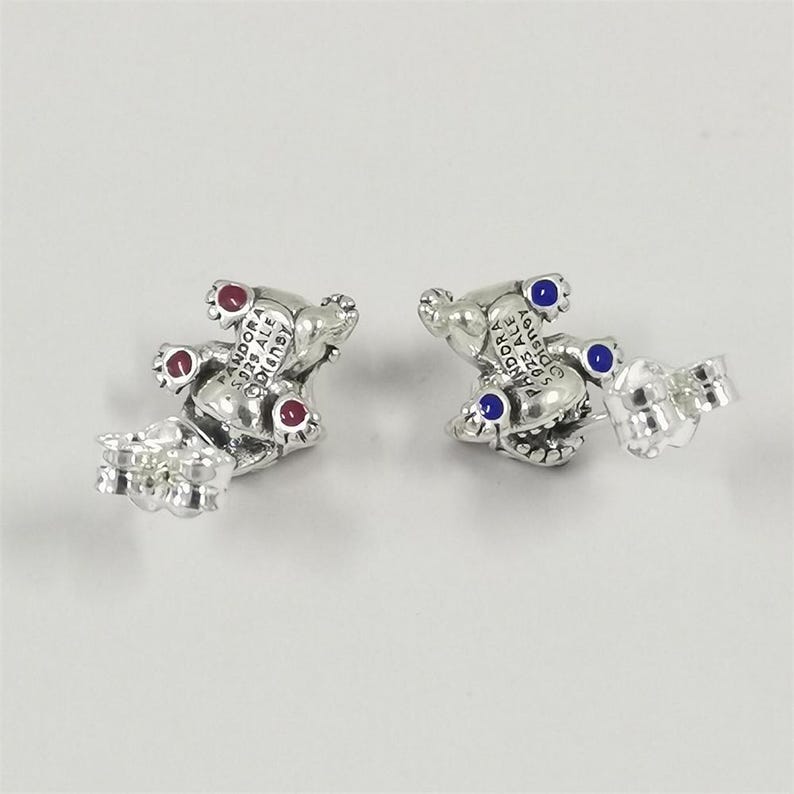 May include: A pair of sterling silver stud earrings shaped like small figures, each featuring the engraved text PANDORA S925 ALE DISNEY. One earring has three red circular accents, while the other earring has three blue circular accents on the feet.