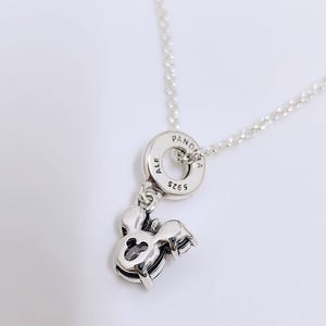 Op de afbeelding: Een zilverkleurige ketting met een ronde hanger waarop de tekst PANDORA, ALE en S925 is gegraveerd. Aan de ring hangt een bedel in de vorm van een gestileerde muizenkop met een kleine uitsparing in het midden en een donkere steen.