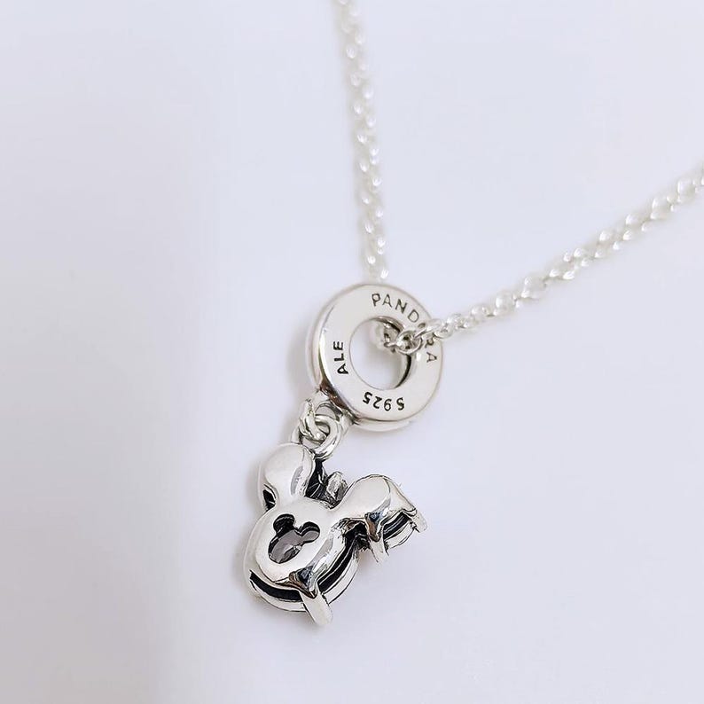 Op de afbeelding: Een zilverkleurige ketting met een ronde hanger waarop de tekst PANDORA, ALE en S925 is gegraveerd. Aan de ring hangt een bedel in de vorm van een gestileerde muizenkop met een kleine uitsparing in het midden en een donkere steen.