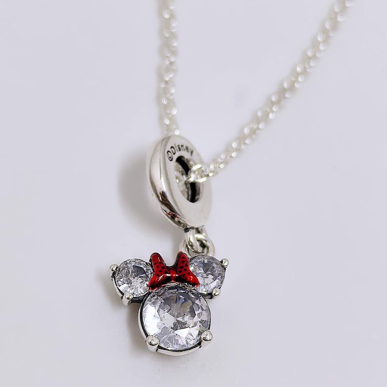 Op de afbeelding: Een zilverkleurige ketting met een hanger in de vorm van een muizenkop, bezet met drie heldere, rondgeslepen zirkonia-stenen. De hanger is voorzien van een kleine rode emaille strik en hangt aan een zilveren ketting met een Disney-oogje.