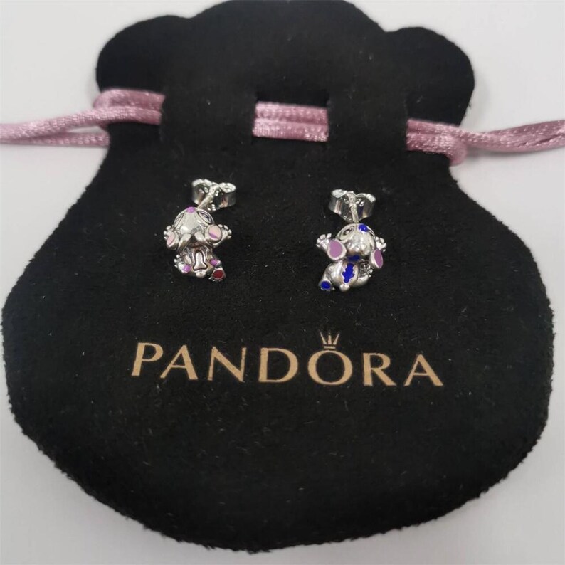 May include: A pair of silver stud earrings featuring a small character design with purple, blue, and red enamel accents. The earrings are displayed on a black velvet pouch with the text PANDORA printed in gold lettering and a pink drawstring.