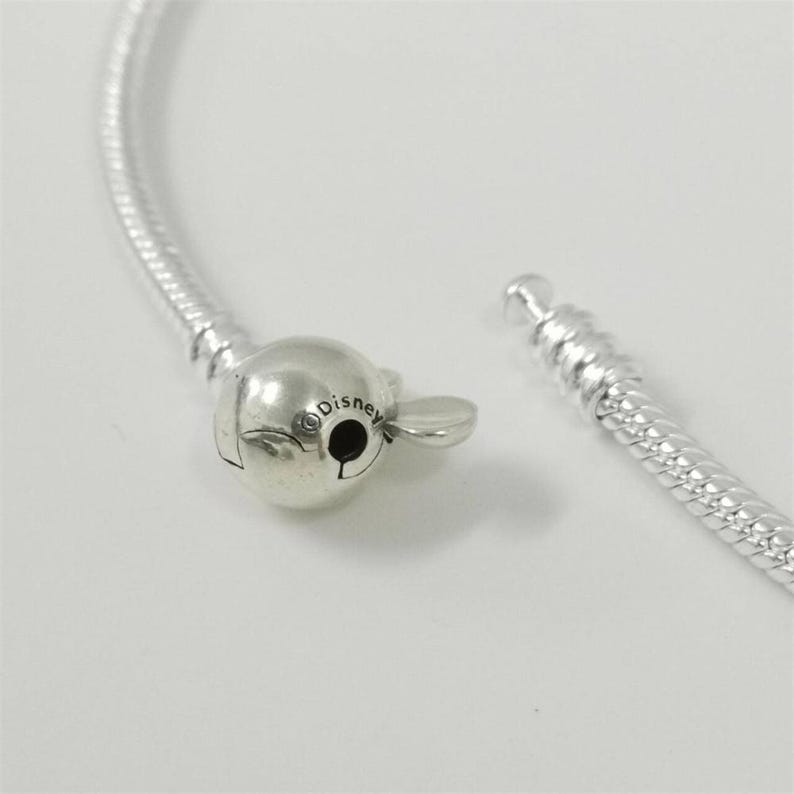 May include: A silver-toned snake chain bracelet featuring a spherical clasp shaped like a Mickey Mouse head. The clasp is engraved with the word Disney and includes a small, flat, rounded tab for opening the mechanism of the jewelry piece.