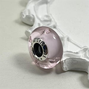 Könnte beinhalten: Eine hellrosa Glasperle mit einem Kern aus silberfarbenem Metall. Die Perle hat eine glatte, abgerundete Form und ist mit "S925" und anderem Text markiert. Die Perle ist ein Charm für ein Armband oder eine Halskette.