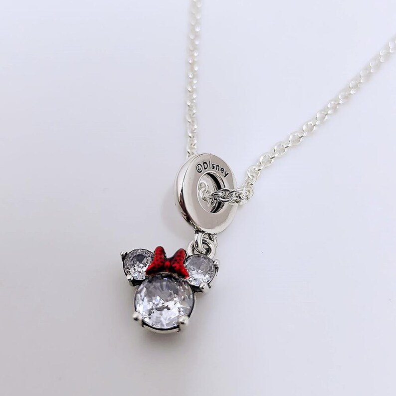Op de afbeelding: Een zilverkleurige ketting met een hanger in de vorm van een muizenkop, voorzien van drie heldere, rondgeslepen stenen en een kleine rode strik met zwarte stippen. De hanger hangt aan een ronde zilveren ring met de inscriptie Disney.