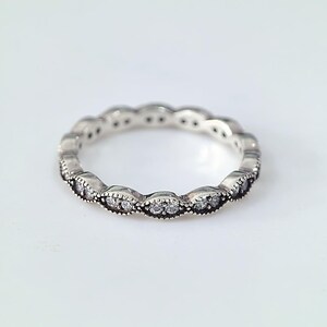 Könnte beinhalten: Ein silberfarbener Ring mit einem sich wiederholenden, marquiseförmigen Muster entlang des Umfangs. Jeder marquiseförmige Abschnitt ist mit einem Perlenrand versehen und enthält zwei kleine, klare, rund geschliffene Steine.