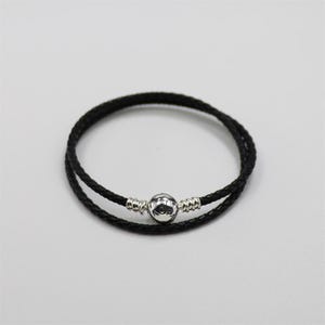 May include: A black braided leather bracelet with a silver clasp. The bracelet is made of two strands of leather and has a round, silver clasp in the center. The bracelet is designed to be worn around the wrist.