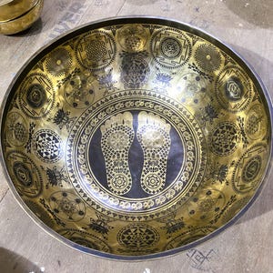 May include: A large, golden singing bowl with intricate etched designs. The bowl features various geometric patterns and symbols, including lotus flowers and the imprint of two feet. The bowl's rim is silver, and the interior is a polished gold color.