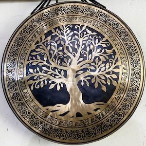 May include: A large, round gong with a brass finish and a dark blue center. The gong features a detailed tree of life design in the center and an ornate border. Black rope is attached for hanging. Ideal for meditation and sound healing.