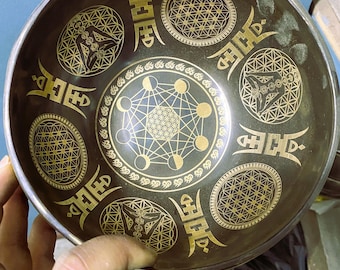 Può includere: Una ciotola tibetana color bronzo con intricati motivi geometrici dorati. La ciotola presenta disegni tra cui il Fiore della Vita, un motivo a forma di stella e forme di luna crescente. La ciotola è tenuta da una mano, mostrando le sue dimensioni e i dettagli.