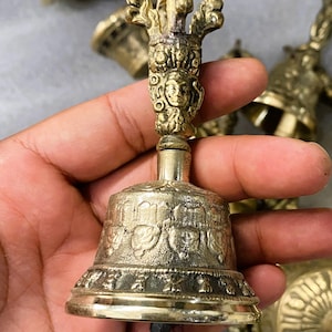 May include: A detailed close-up of a golden bell with intricate carvings. The bell features a detailed handle with a face and ornate designs. Other bells are visible in the background. The bell is held in a hand.