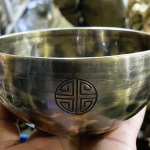Pode incluir: Uma tigela de canto de latão com um design decorativo gravado. A tigela tem uma forma arredondada e uma superfície lisa e polida. O design gravado é um padrão simétrico dentro de um círculo. A tigela é segurada em uma mão.