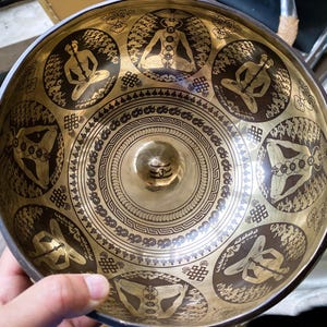 Puede incluir: Un cuenco tibetano de latón con intrincados diseños grabados. El cuenco presenta medallones circulares con figuras en posturas de meditación y patrones detallados. El interior tiene un área central elevada y es sostenido por una mano.
