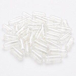 May include: A collection of clear, empty gelatin capsules, scattered on a white surface. The capsules are cylindrical with rounded ends, designed for filling with powders or supplements. The transparent material allows visibility of the contents.