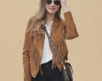 Pode incluir: Casaco de camurça castanho com franjas, combinado com calças pretas e uma pequena bolsa castanha com uma alça estampada. O casaco tem um estilo western clássico, e o look é completado com óculos de sol aviador.