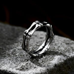 May include: A silver-toned ring with a skeletal bone design. The ring features multiple bone-shaped elements arranged to form a band. The ring is displayed on a textured gray surface.