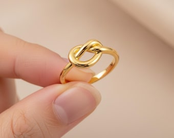 Puede incluir: Un anillo dorado con un diseño de nudo. El anillo está sostenido por una mano, mostrando su superficie pulida y los intrincados detalles del nudo. La banda del anillo es lisa y redondeada, siendo el nudo el punto focal del diseño.