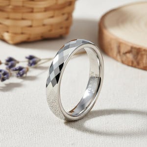 Può includere: Un anello color argento con un design sfaccettato sulla superficie superiore. L'anello ha una fascia liscia e lucida e una superficie interna strutturata. L'anello è mostrato su una superficie bianca con uno sfondo sfocato.