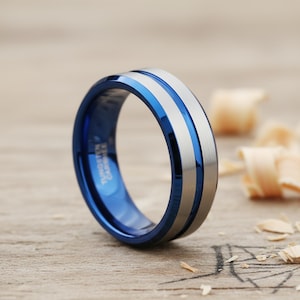 Op de afbeelding: Een blauwe en zilveren wolfraamcarbide ring. De ring heeft een gepolijste blauwe binnenkant en een geborstelde zilveren band met een centrale blauwe streep. De ring staat op een houten achtergrond.