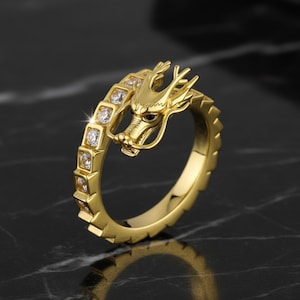 May include: A gold ring with a dragon motif. The dragon's head is prominent, and the band incorporates a series of square-set, clear stones. The ring has a detailed, textured finish.
