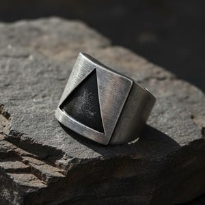 May include: A silver ring with a geometric design. The ring has a dark triangle set within a rectangular frame. The metal has a textured, brushed finish, giving it a vintage or industrial look. The ring is displayed on a grey, textured surface.