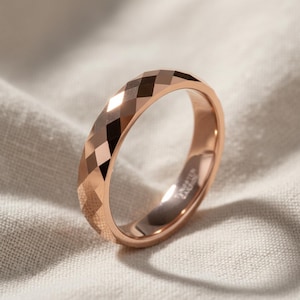Peut inclure: Bague en or rose avec un design &agrave; facettes. La bague pr&eacute;sente un motif g&eacute;om&eacute;trique de losanges autour de l'anneau. La bague est pr&eacute;sent&eacute;e sur un fond en tissu de couleur neutre.
