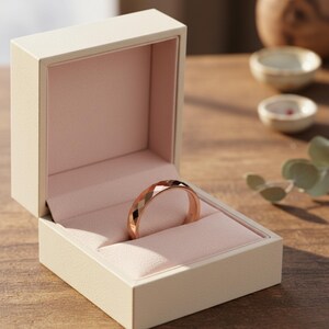 Peut inclure: Une bague en or rose avec un design g&eacute;om&eacute;trique &agrave; facettes repose dans un &eacute;crin &agrave; bijoux cr&egrave;me ouvert avec un int&eacute;rieur rose. La bague est le point central, mettant en valeur sa surface polie et sa coupe complexe. La bo&icirc;te est sur une surface en bois.