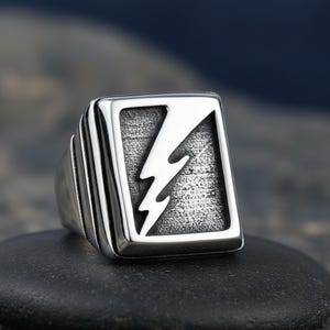 May include: A silver-colored ring with a lightning bolt design. The ring features a square face with a textured background and a white lightning bolt. The band has a ribbed design. The ring is displayed on a black stone.