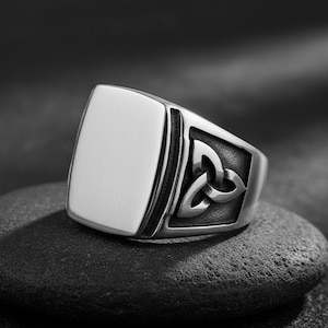Op de afbeelding: Een zilverkleurige ring met een rechthoekig oppervlak en een Keltisch knoopmotief. De gepolijste ring contrasteert met de donkere, getextureerde achtergrond, waardoor de details en de glans van het metaal opvallen.
