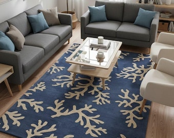 May include: A living room scene featuring a navy blue area rug with a coral reef design in beige. The room includes two gray sofas, accent pillows, a glass-top coffee table, and two white armchairs. A large potted plant adds greenery.
