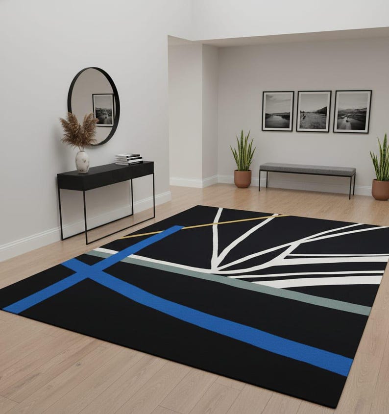 May include: A black rectangular area rug with a geometric design. The rug features a bold blue stripe, white lines, and accents of yellow and gray. The rug is placed on a light wood floor in a room with neutral walls.