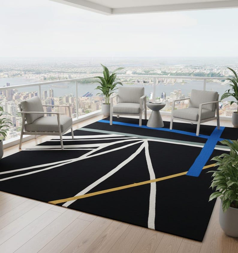 May include: Black outdoor rug with a geometric design featuring white, yellow, and blue lines. The rug is placed on a wooden deck, with two gray chairs and a small table. Potted plants and a city skyline are visible in the background.