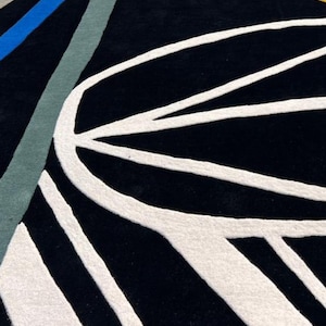 May include: A black rug with a white, abstract design. The rug features curved and straight white lines against a black background. Accents of blue, gray, and yellow-orange are visible in the upper left corner.