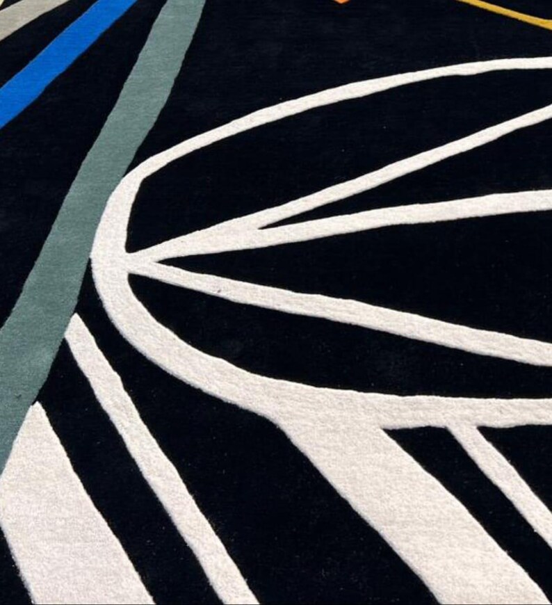 May include: A black rug with a white, abstract design. The rug features curved and straight white lines against a black background. Accents of blue, gray, and yellow-orange are visible in the upper left corner.